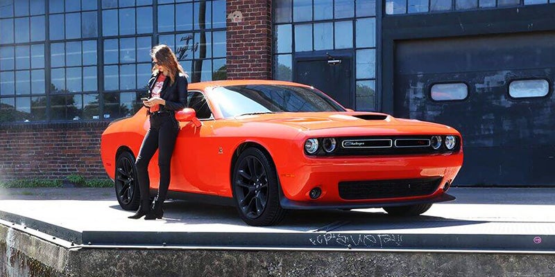 Woman leaning on a bright orange Dodge Challenger, looking at her phone, in front of an industrial building.