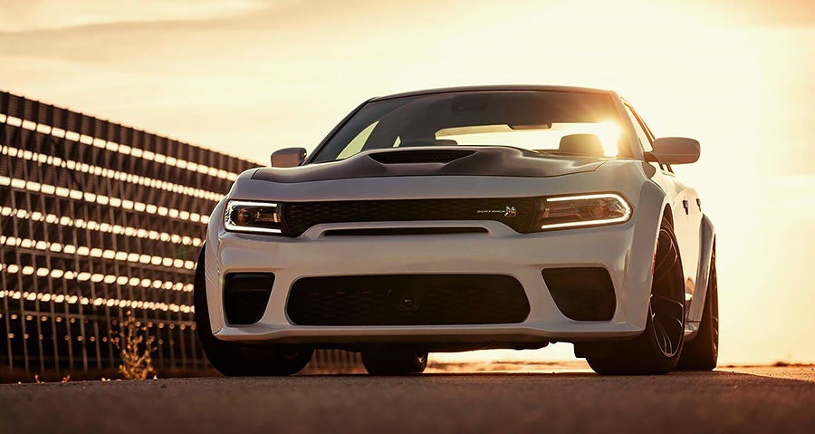 White Dodge Charger Hellcat from the front, black hood, glowing headlights, sun shining brightly from behind.