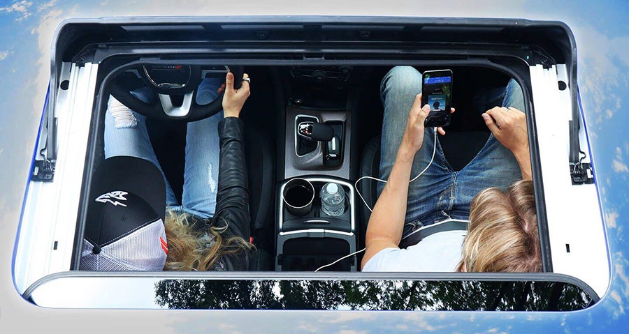 Overhead view through a car sunroof of two people with legs up; one holds the steering wheel, the other a smartphone.