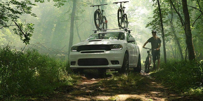 White SUV with two mountain bikes on its roof rack on a dirt path in a misty forest, with a man and bike beside it.