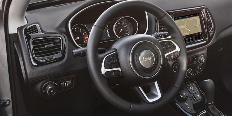 Car interior showing a black steering wheel with a Jeep logo, dashboard, gauges, and an infotainment screen with a map.