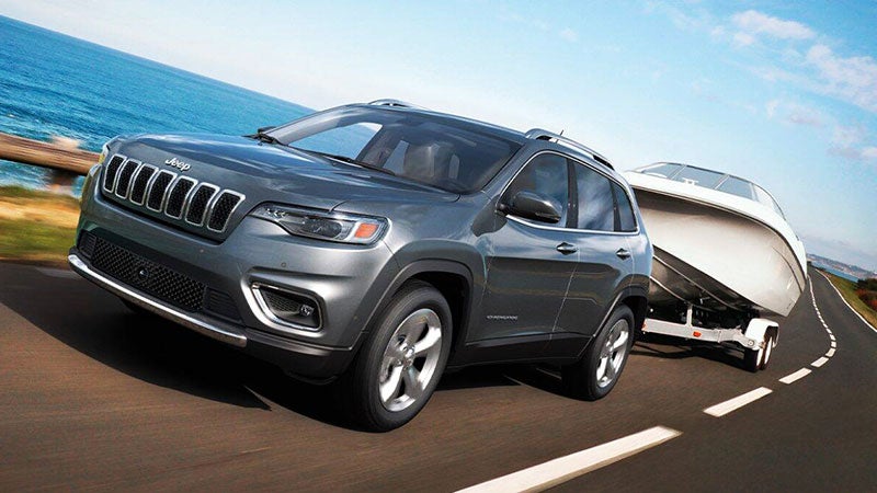 Grey Jeep Cherokee SUV towing a white boat on a trailer, driving on a coastal road with the ocean in the background.
