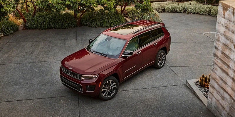 Dark red SUV with a panoramic roof and roof rails, parked on a paved driveway surrounded by greenery and a stone wall.