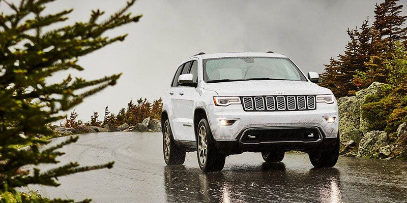 White Jeep Grand Cherokee on a wet road with trees and rocks under an overcast sky.