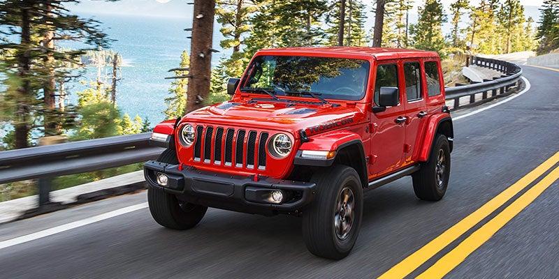 Red Jeep Wrangler driving on a winding mountain road with a large body of water and pine trees in the background.