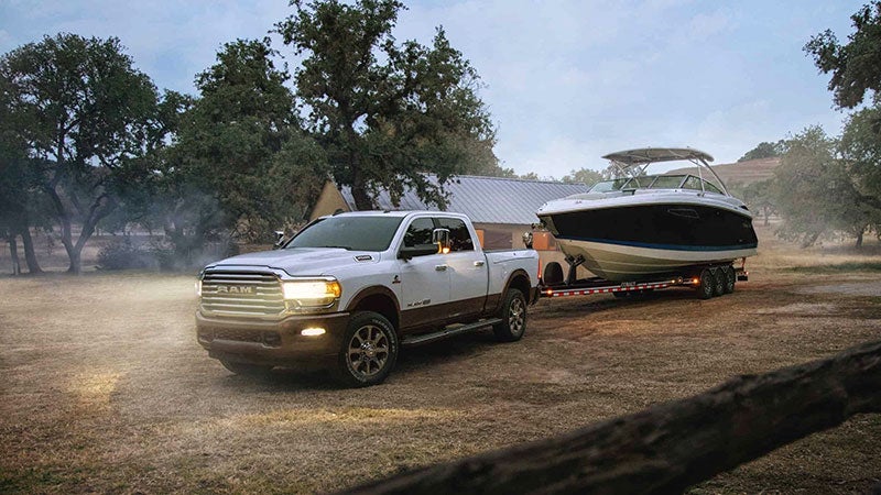 Two-tone RAM pickup truck with headlights on, towing a white and blue boat on a trailer in a rural, grassy area.