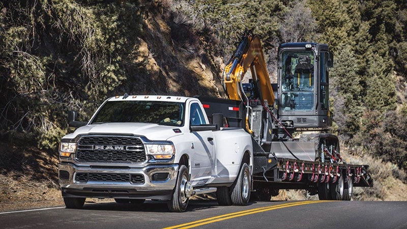 White Ram pickup truck towing a trailer with a yellow and black excavator on a winding road.