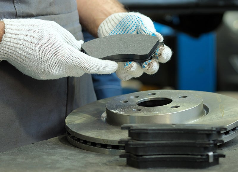 Hands in white work gloves hold a new brake pad over a shiny brake rotor and other new pads.